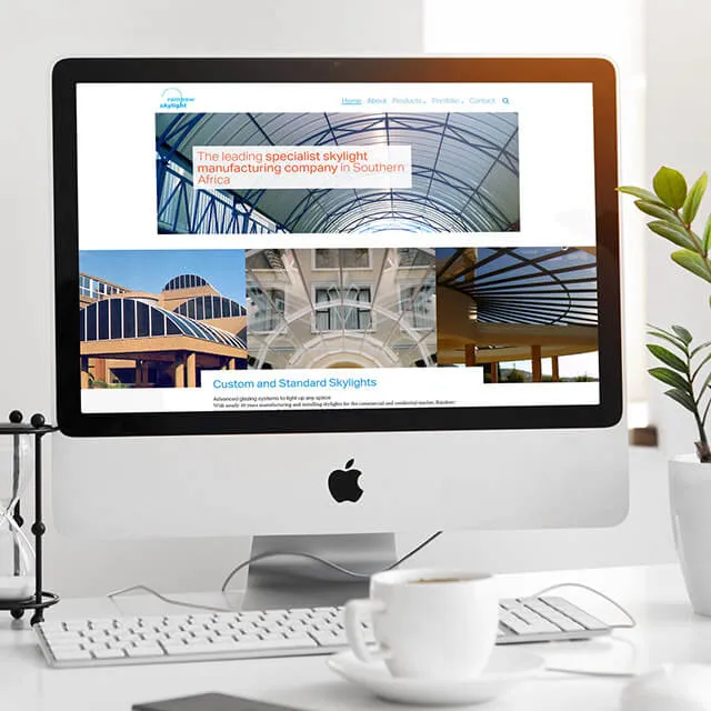 The Rainbow Skylight home page on an iMac on a white desk with a coffee cup and a plant. A wide photograph of a barrel-vaulted glass roof fills the hero, with the line "The leading specialist skylight manufacturing company in Southern Africa" set over a translucent white panel.